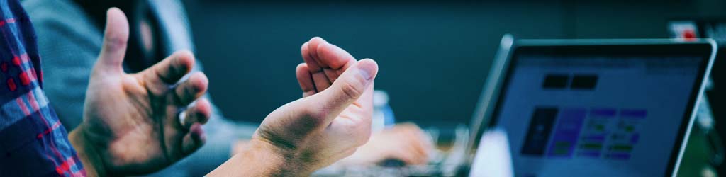 A picture of hands gesturing in discussion near an open laptop with a blurred screen.