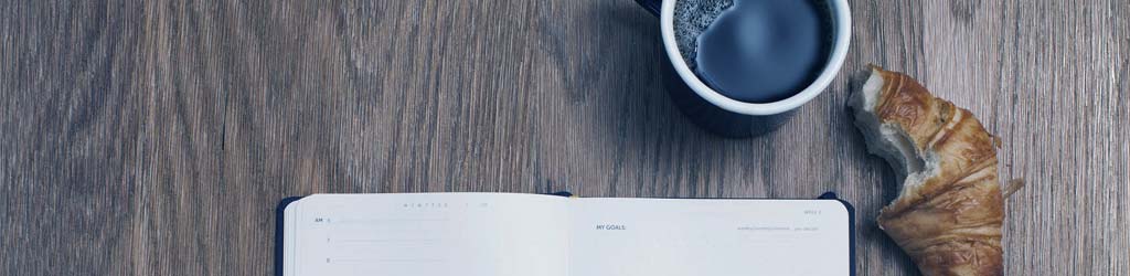Photograph of a notebook on a desk. A croissant and a coffee cup sit next to it.